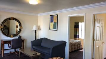 A hotel room with a desk, a couch, a bed, a mirror, a lamp, and a door leading to another room.