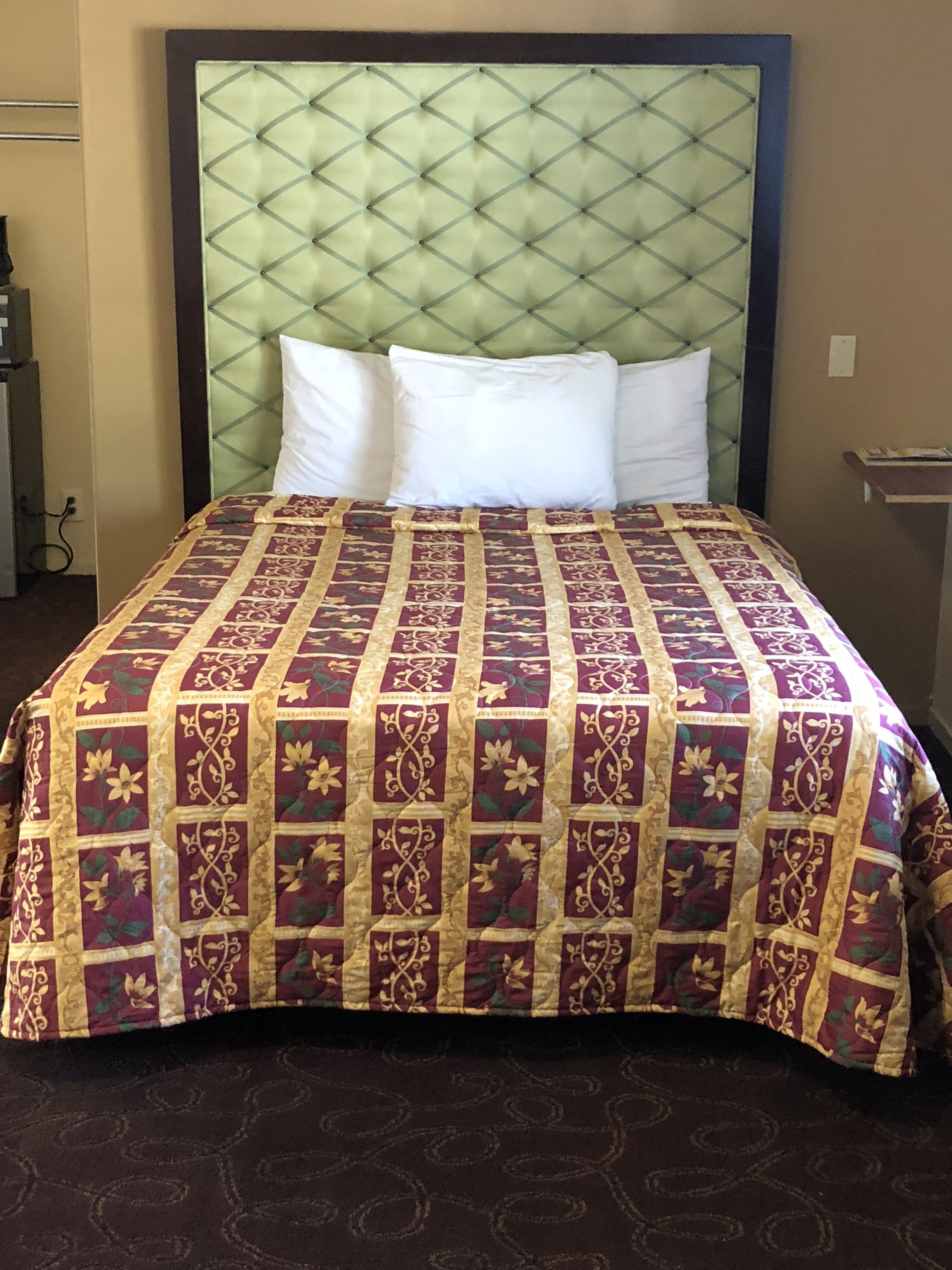 Una cama con colcha estampada, dos almohadas blancas, cabecera capitonada y alfombra marrón oscuro.