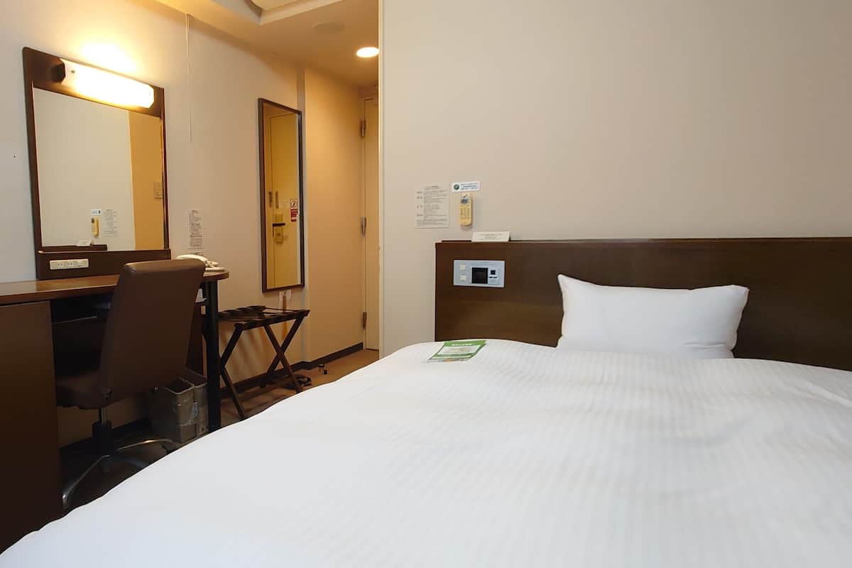 Quarto de hotel com cama, escrivaninha, cadeira, espelho e termostato fixado na parede.