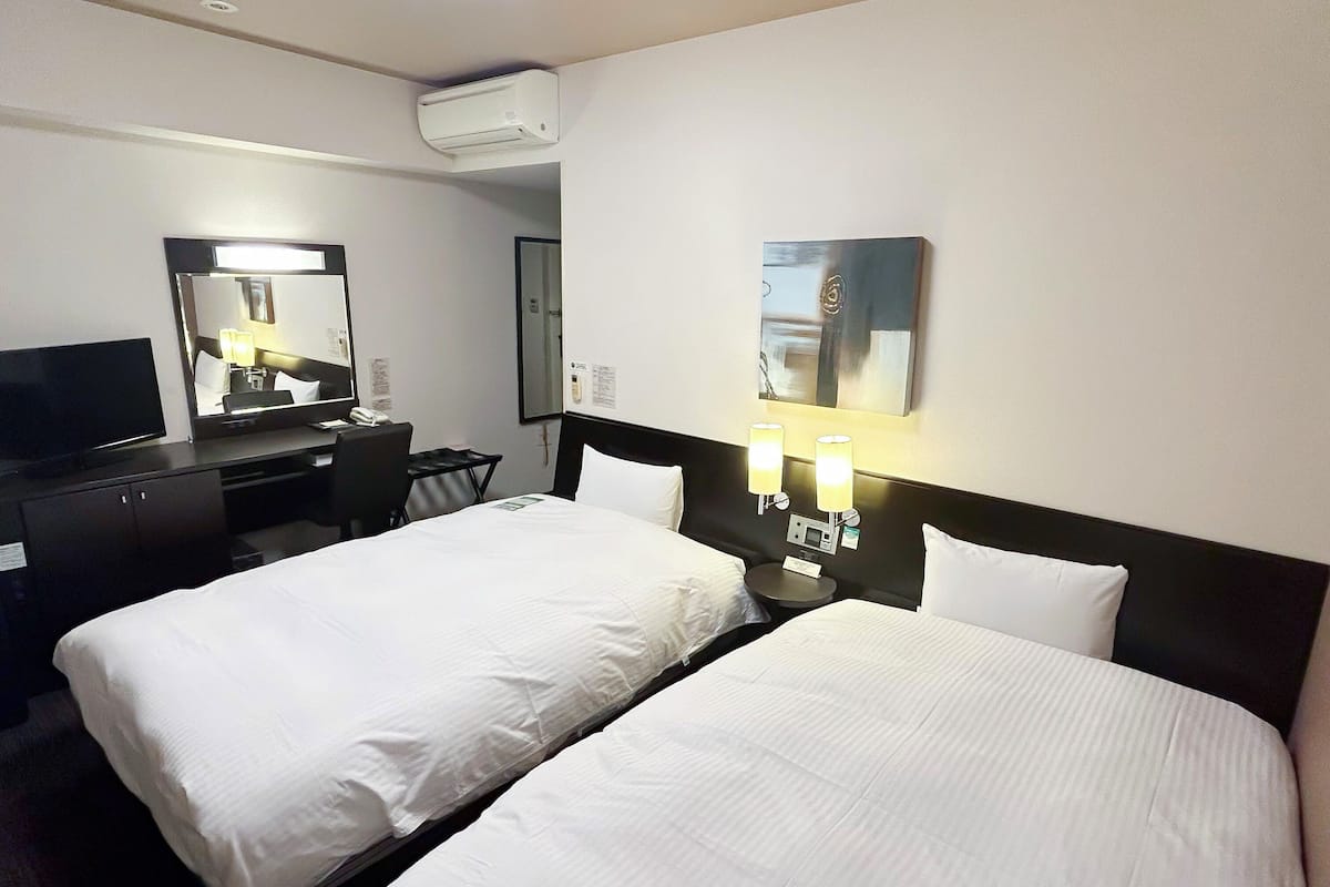 Quarto de hotel com duas camas, uma escrivaninha com cadeira, televisão, ar condicionado e luminárias fixadas na parede.