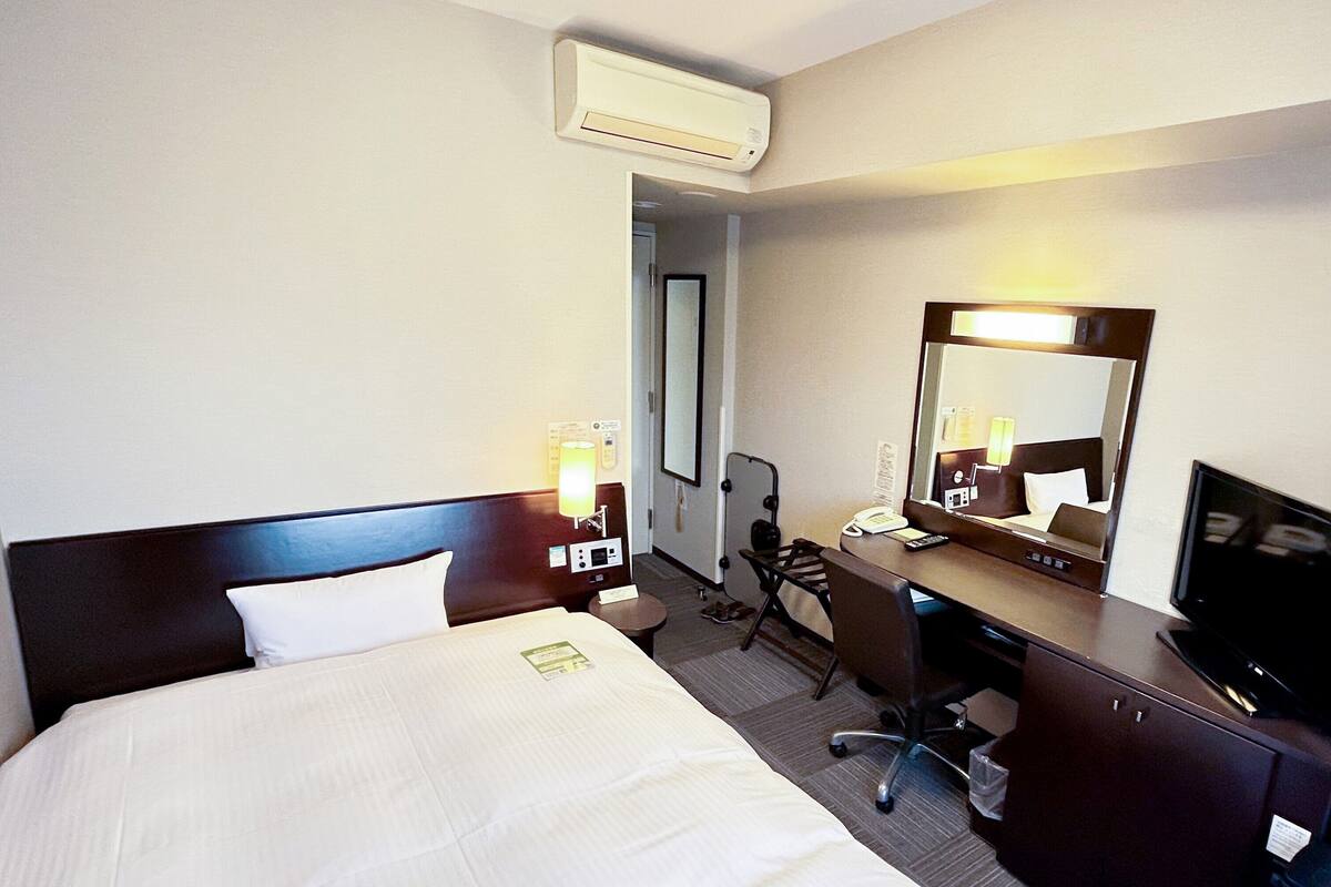 Quarto de hotel com uma cama grande, uma escrivaninha, uma cadeira, televisão, espelho e ar condicionado.