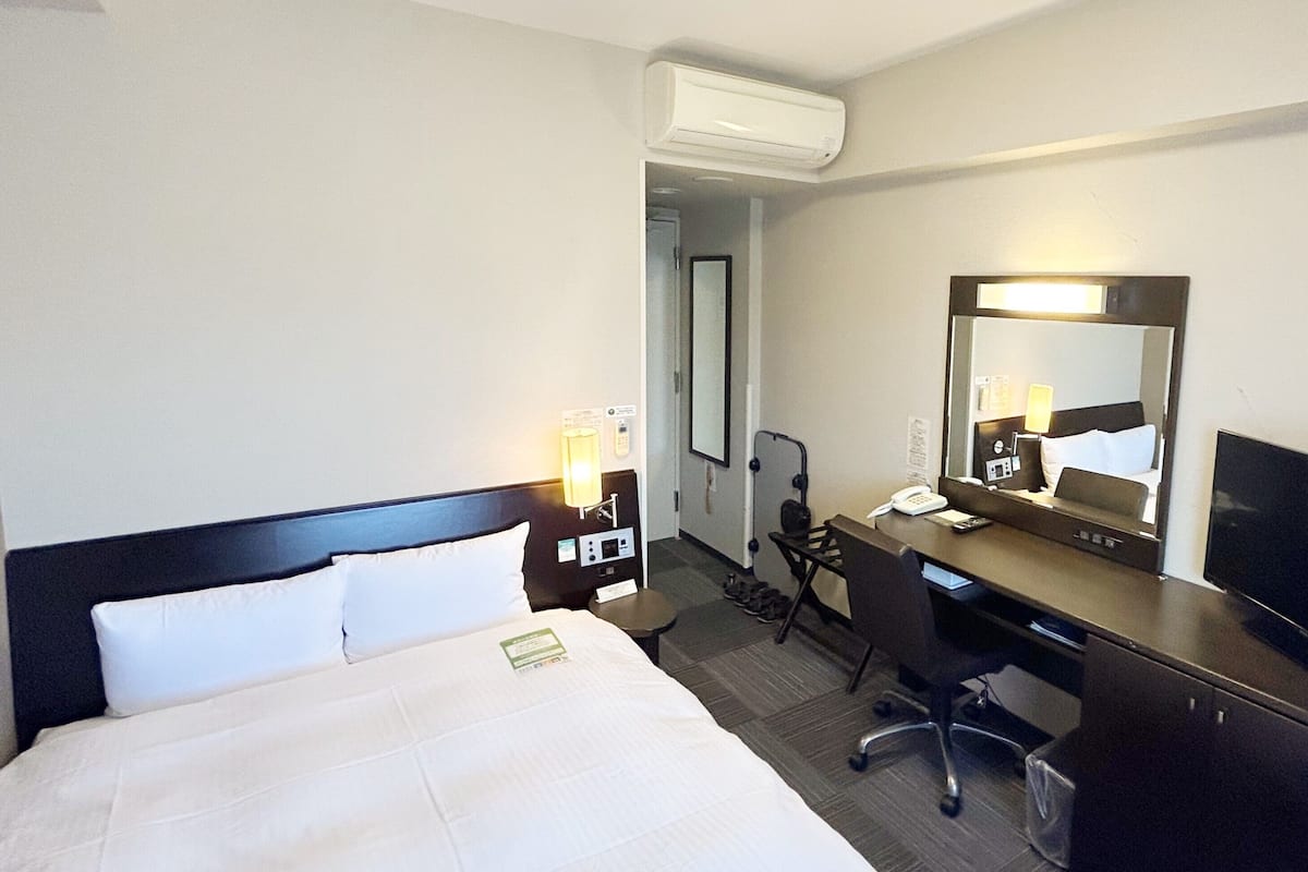 Quarto de hotel com cama, escrivaninha, cadeira e um espelho grande.