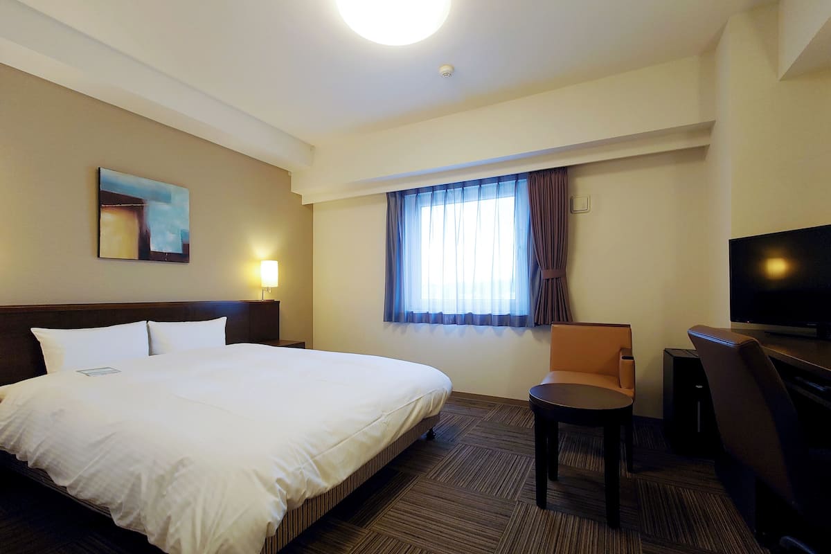 Quarto de hotel com uma cama grande, uma escrivaninha com cadeira, televisão e janela com cortinas.