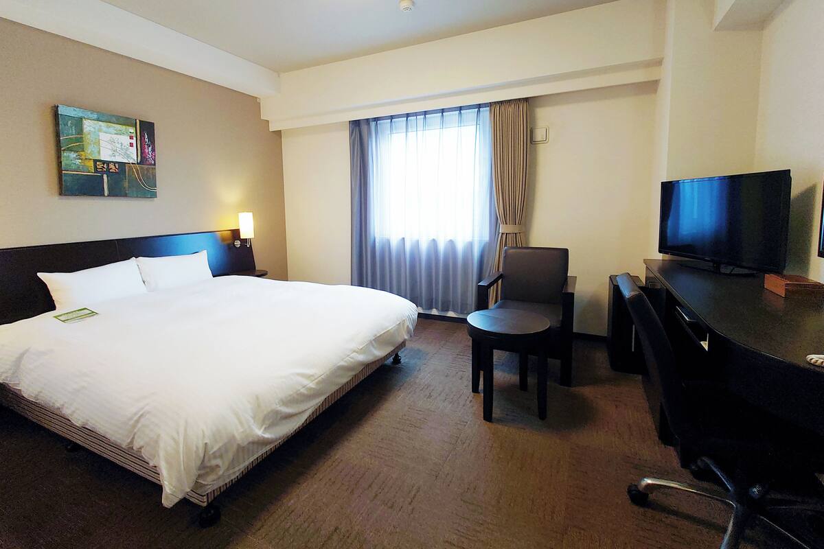 Quarto de hotel com uma cama grande, uma escrivaninha com cadeira, televisão e janela com cortinas.
