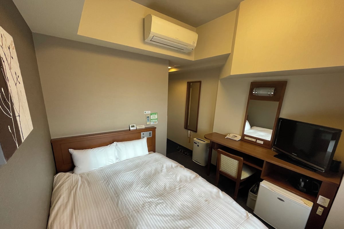 A hotel room with a bed, a desk with a chair, a television, a mini-fridge, and an air conditioning unit.