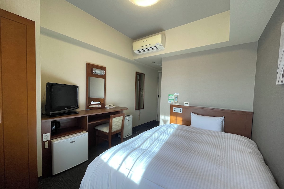A hotel room with a bed, a desk with a computer, a chair, a small refrigerator, and an air conditioning unit.