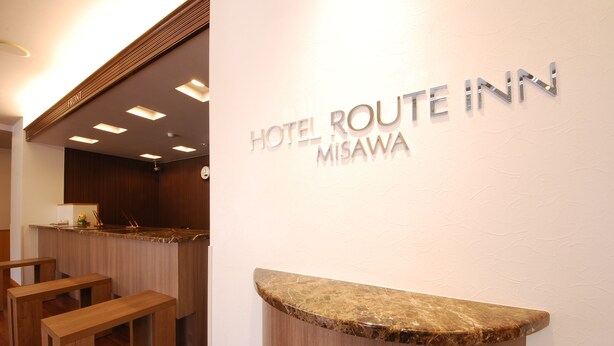 Un comptoir d'accueil avec une enseigne indiquant « HÔTEL ROUTE INN MISAWA » et des bancs en bois en arrière-plan.