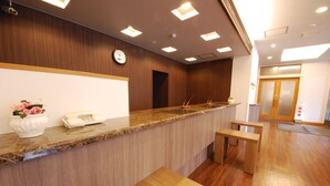 Un espace d’accueil avec un comptoir en bois, une horloge et un téléphone. On y trouve deux bancs en bois et une plante en pot ornée de fleurs roses.