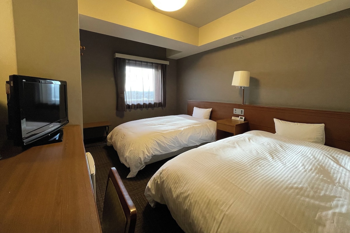 A hotel room with two beds, a desk, a television, a lamp, and a window with curtains.