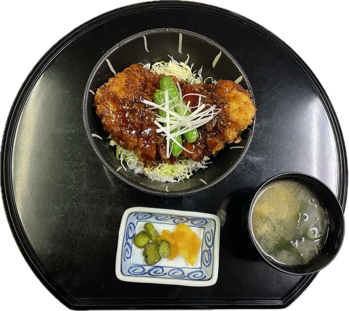 Un plato que incluye un alimento empanizado y frito, posiblemente pollo, sobre una cama de col rallada, acompañado de un tazón pequeño de sopa y un plato con vegetales encurtidos.