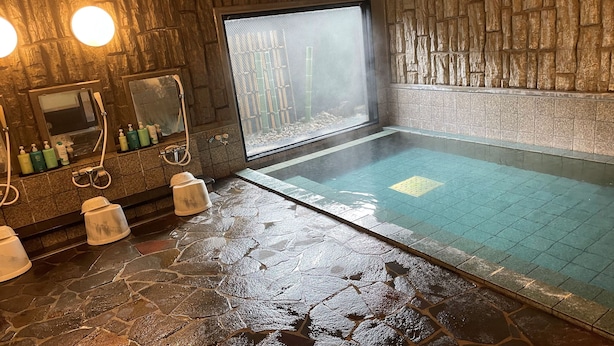 A stone-floored bathroom with a large bathtub, a window with a view of bamboo, and toiletries on the counter.