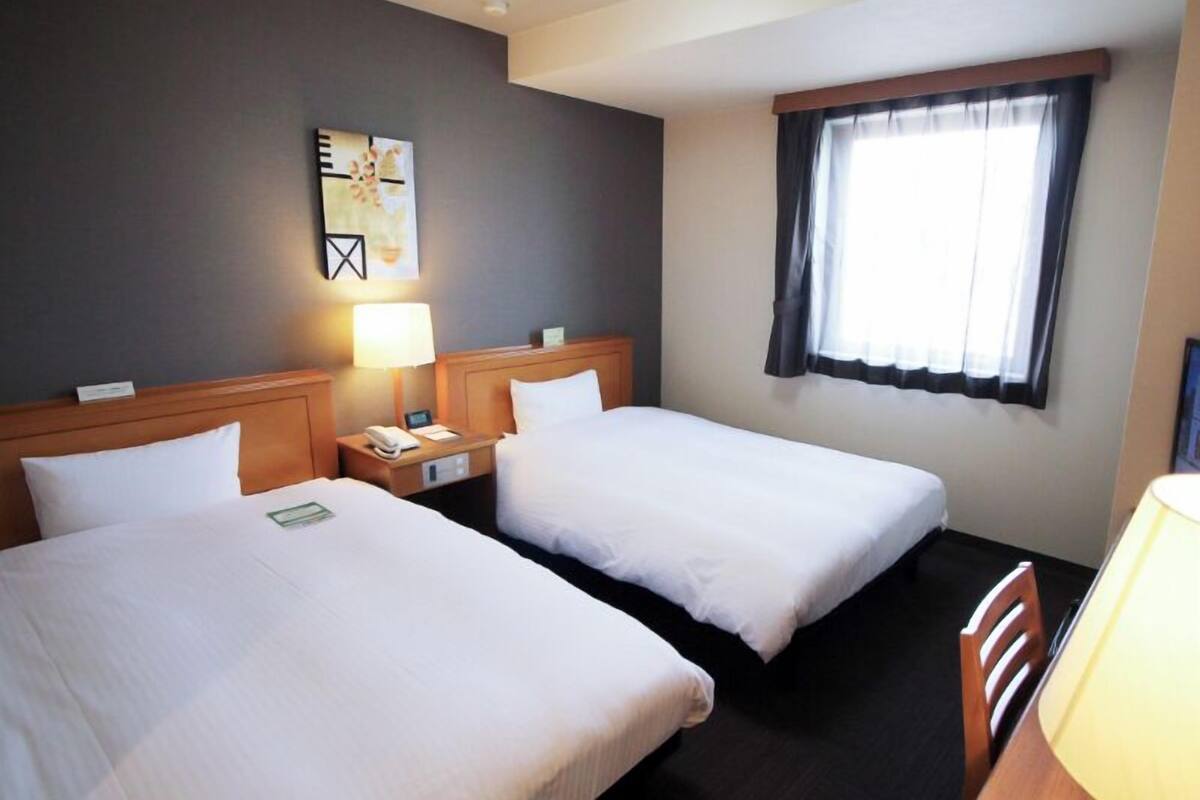 Hotellihuone, jossa on kaksi erillistä sänkyä, yöpöytä lampun kanssa, taulu seinällä ja ikkuna, jossa on verhot.
