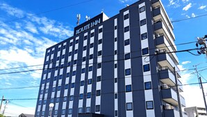 A multi-story building with the sign "ROUTE INN" indicating it is a hotel. The building has a modern architectural style with a combination of dark and light panels. There are several windows visible, some with curtains, and balconies on the upper floors.