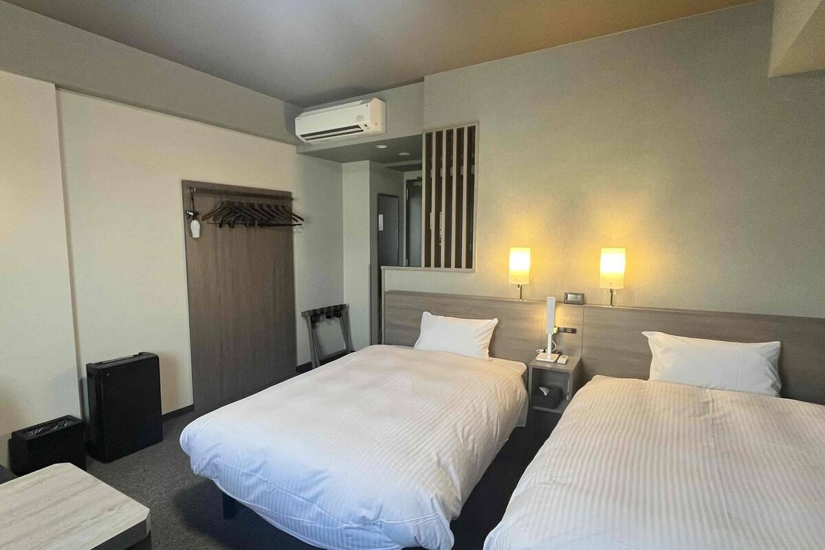 A hotel room with two beds, a desk, a chair, an air conditioning unit, and two wall-mounted lamps.