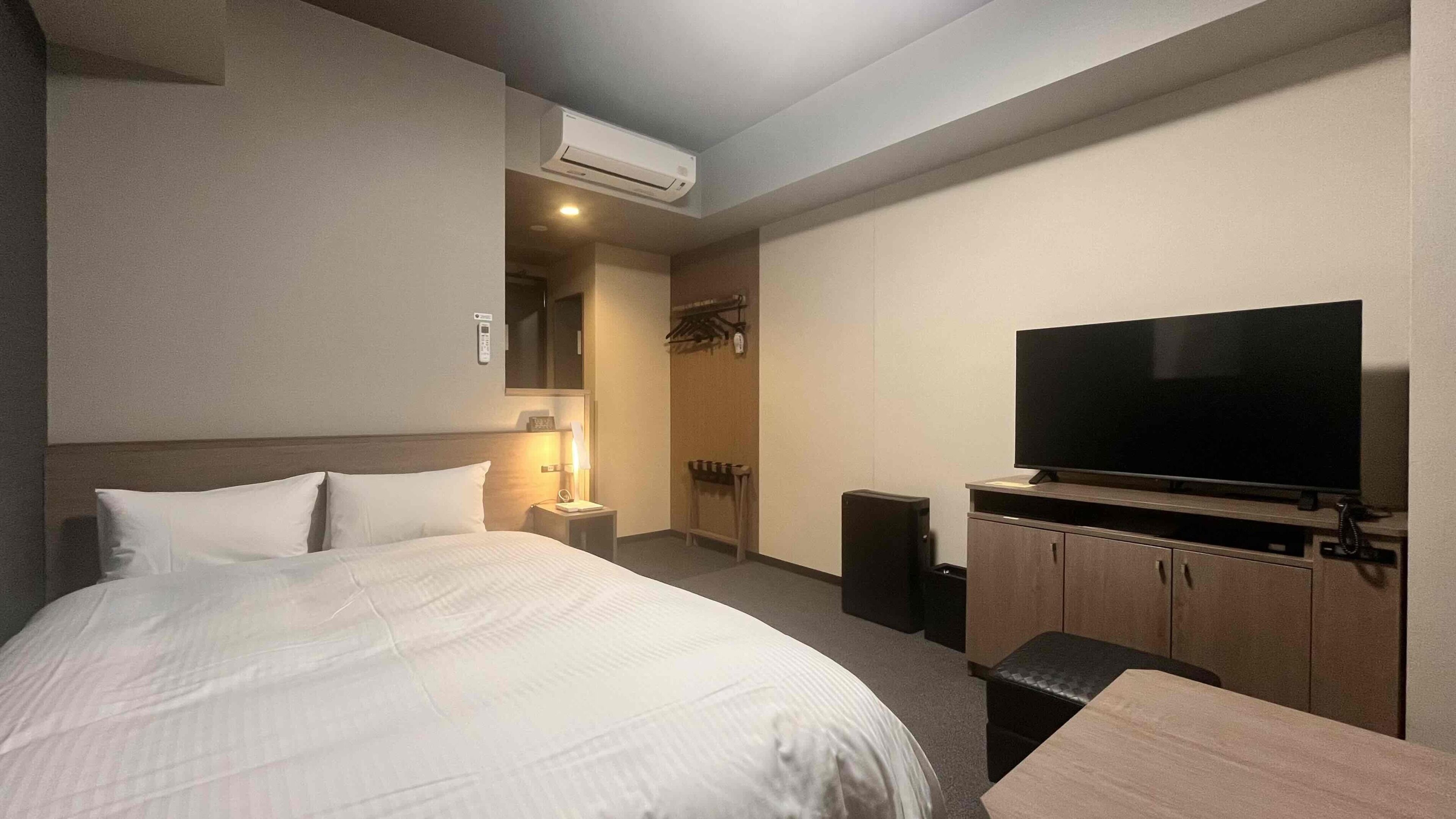 A hotel room with a bed, a television, a bedside table, a lamp, and an air conditioning unit.