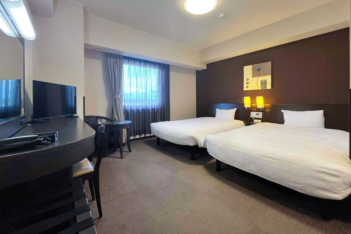 A hotel room with two beds, a desk, a TV, and a window with curtains.