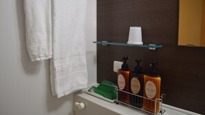 A bathroom with a white towel hanging on the wall, a glass shelf with a cup and three bottles of body soap.