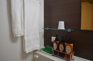A bathroom with a white towel hanging on the wall, a glass shelf with a cup and three bottles of body soap.