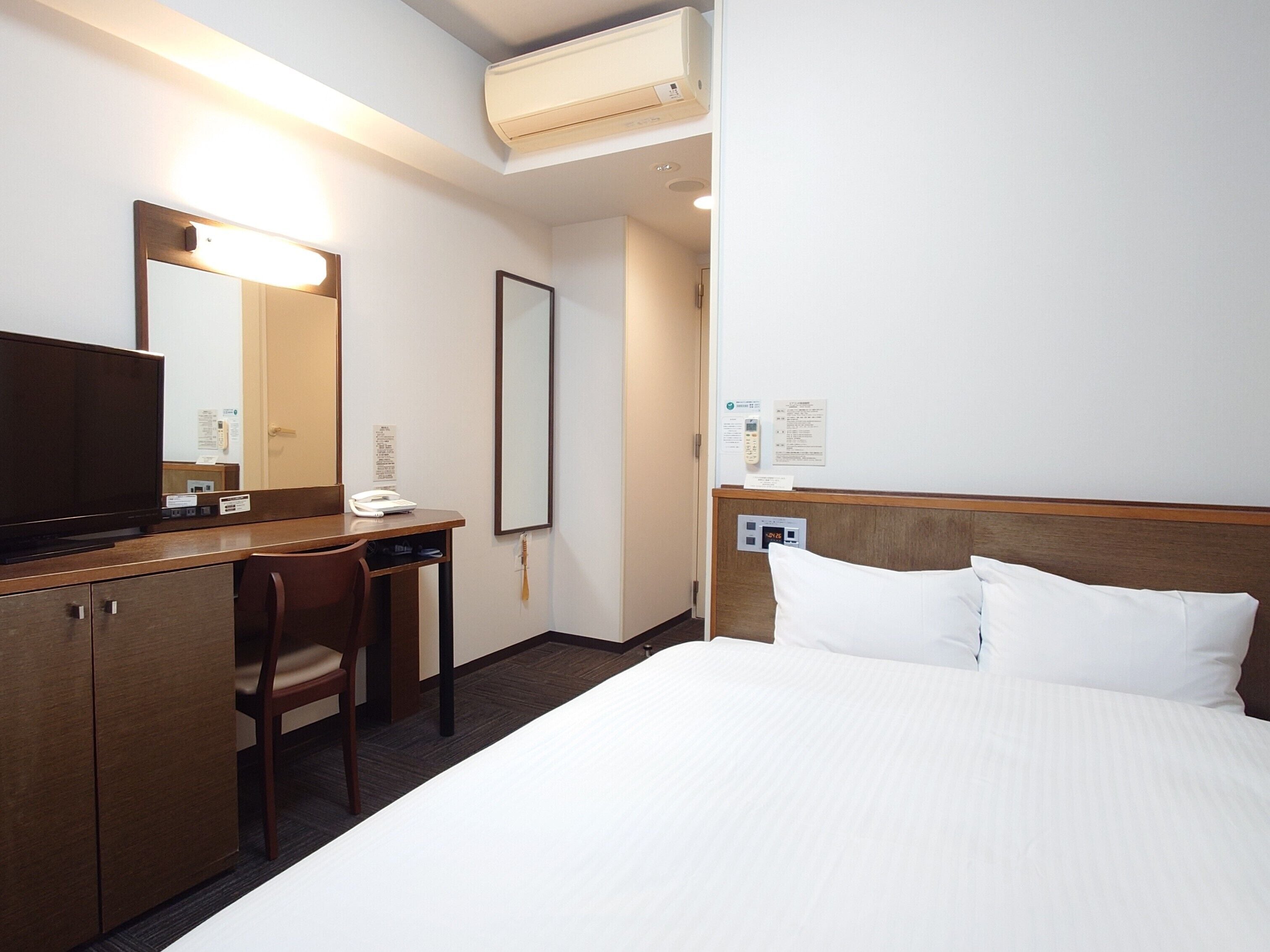 A hotel room with a bed, a desk, a chair, a television, a mirror, and a wall-mounted phone.