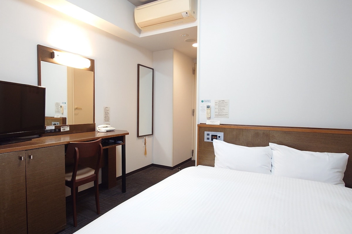 A hotel room with a bed, a desk, a chair, a television, a mirror, and a wall-mounted phone.