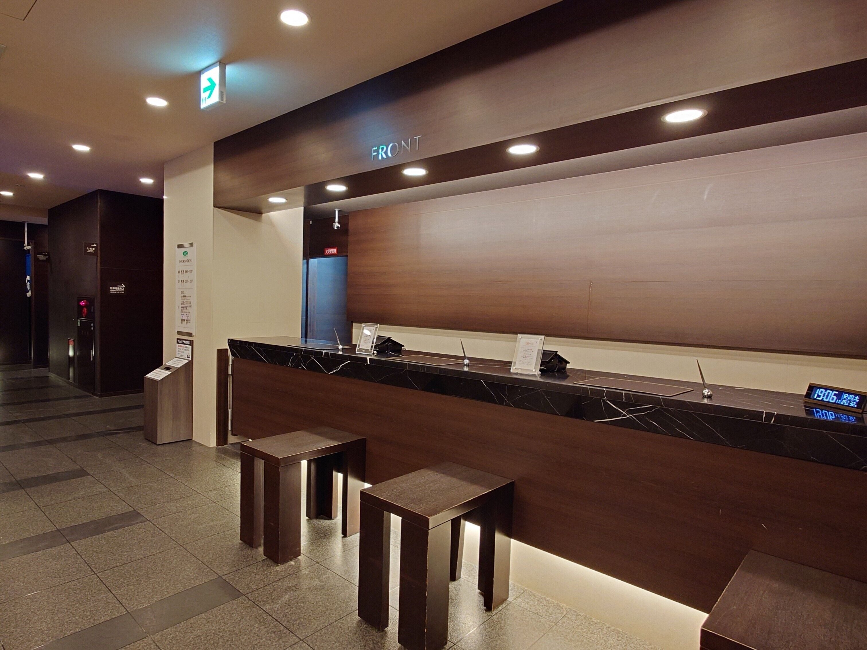 A reception desk with a digital clock displaying 19:06, a sign indicating the front area, and a blue elevator door in the background.