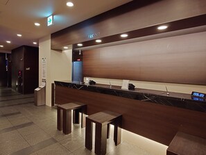 A reception desk with a digital clock displaying 19:06, a sign indicating the front area, and a blue elevator door in the background. - Hotel Route - Inn Yurihonjyo (Yurihonjo)