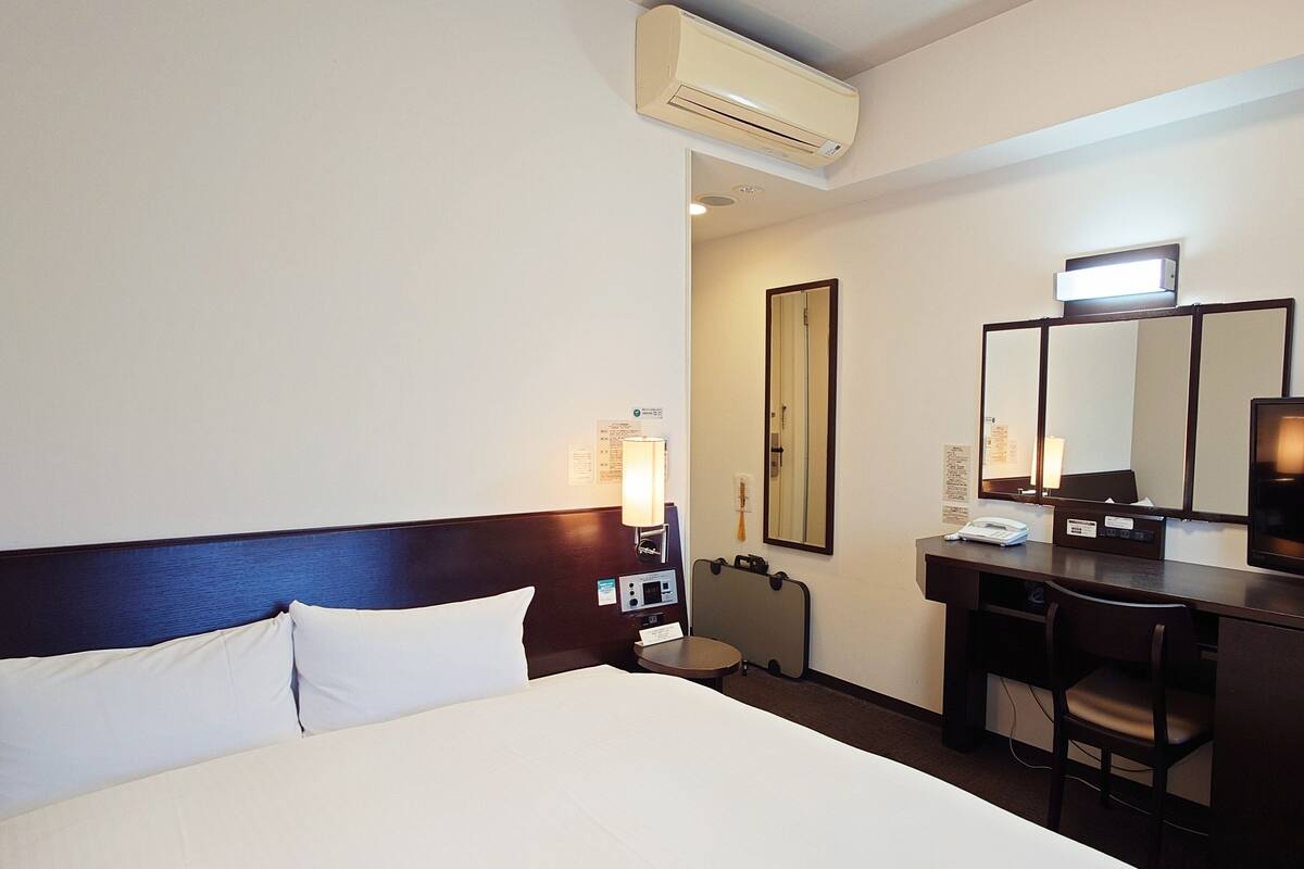 A hotel room with a bed, a desk, a chair, a desk lamp, a mirror, an air conditioning unit, and a wall-mounted TV.
