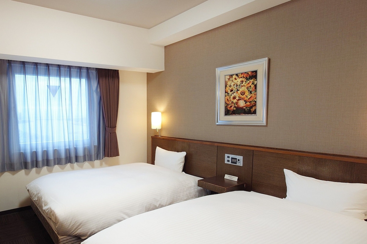 A hotel room with two beds, a bedside table, a lamp, a painting on the wall, and a window with curtains.