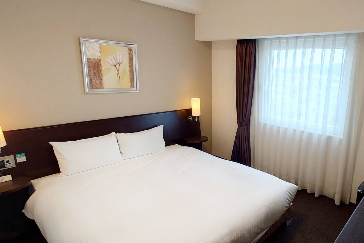 A hotel room with a large bed, two bedside lamps, a nightstand, a framed picture on the wall, and a window with curtains.