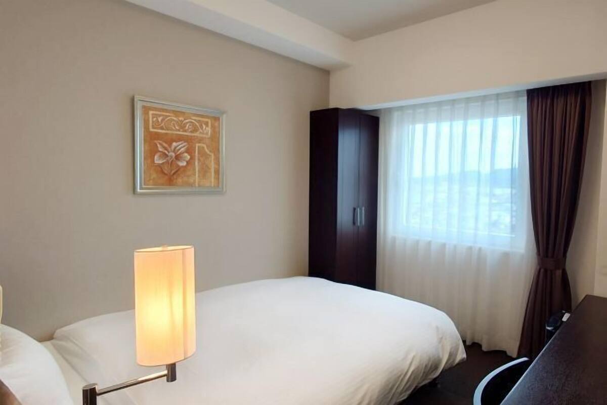A hotel room with a bed, a desk, a lamp, a wardrobe, and a window with curtains.