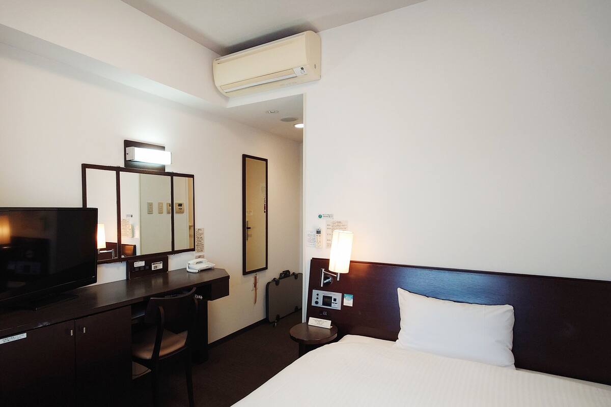 A hotel room with a bed, a desk, a chair, a television, a mirror, a nightstand, and an air conditioning unit.