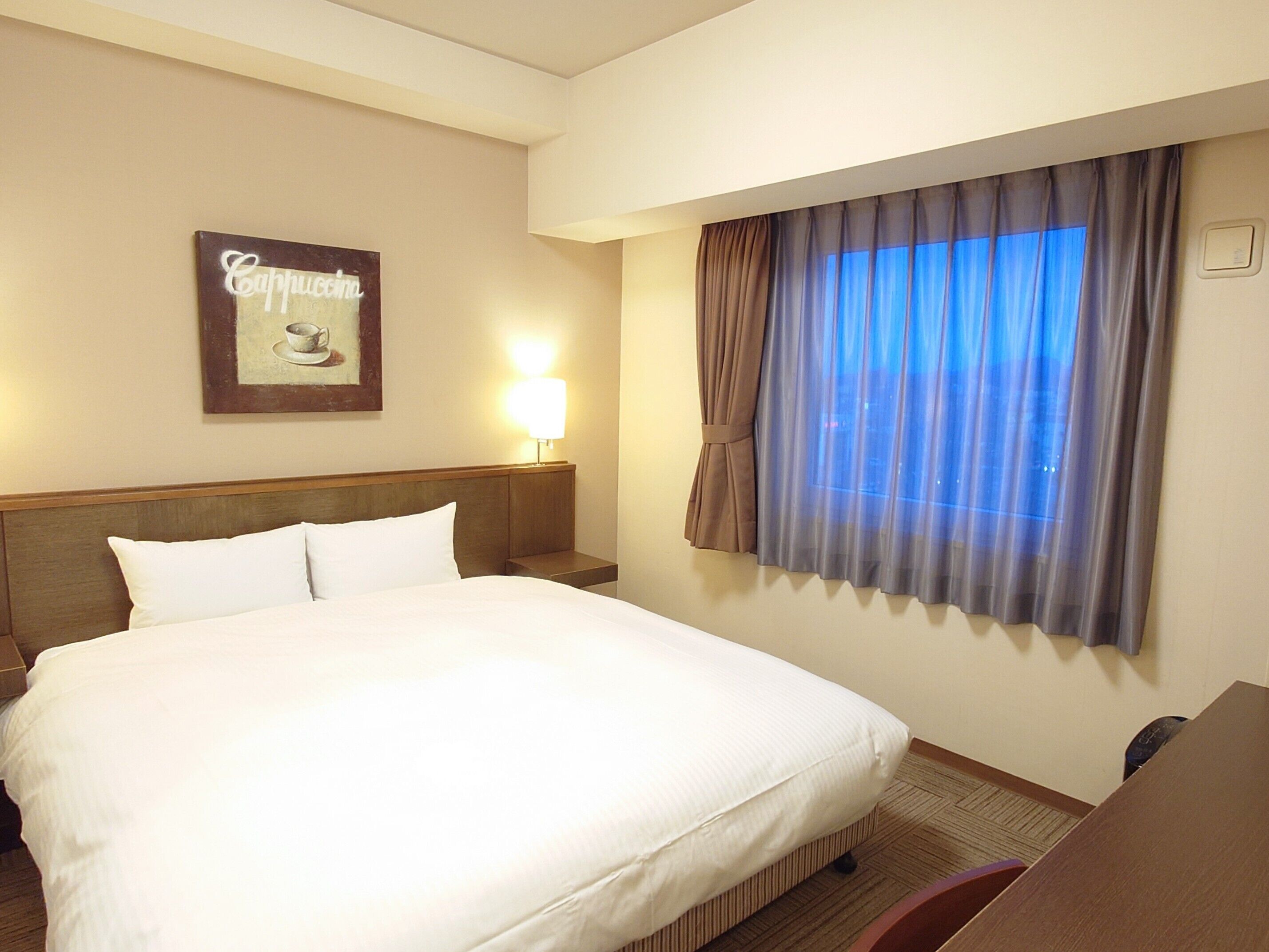 A hotel room with a large bed, a nightstand with a lamp, a window with curtains, and a wall-mounted sign.