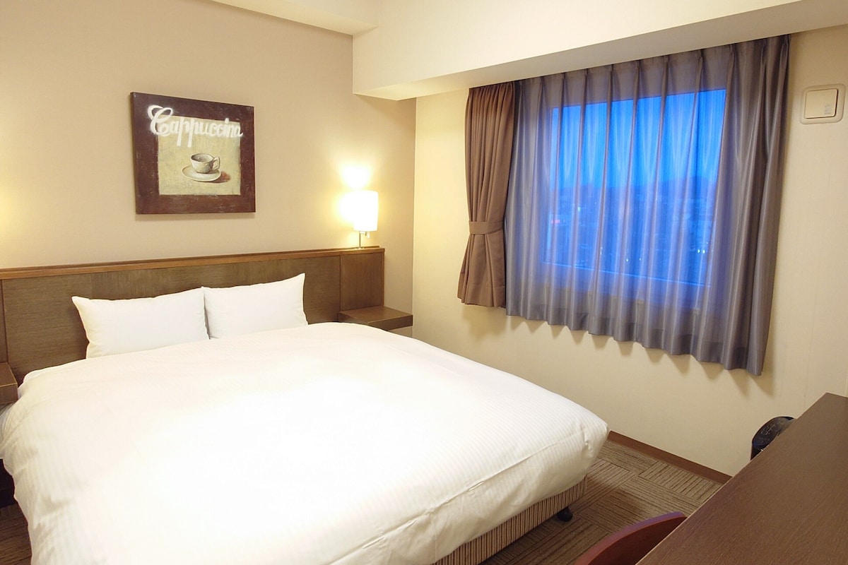 A hotel room with a large bed, a nightstand with a lamp, a window with curtains, and a wall-mounted sign.