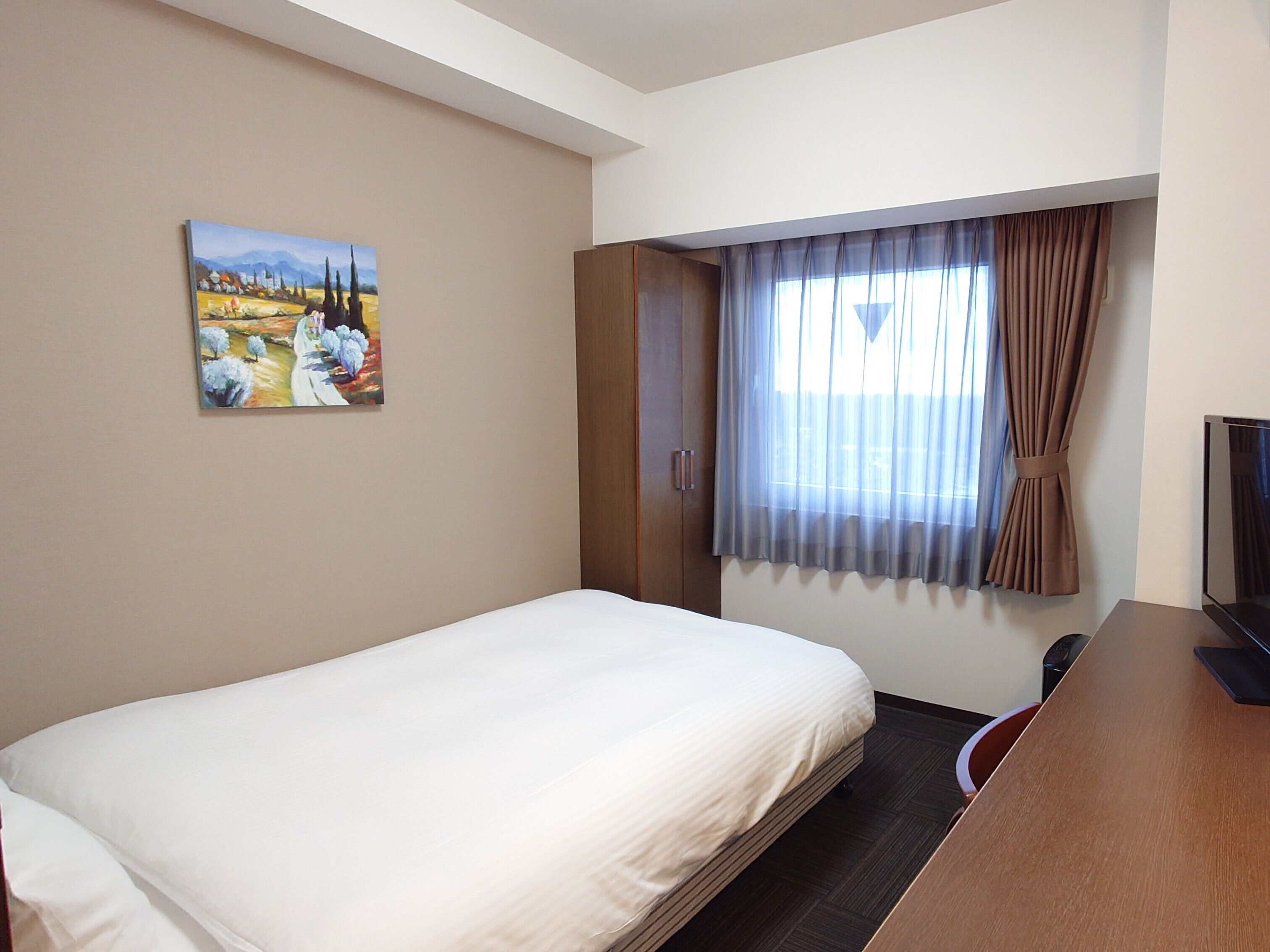 A hotel room with a bed, a painting on the wall, a window with curtains, and a desk with a television.