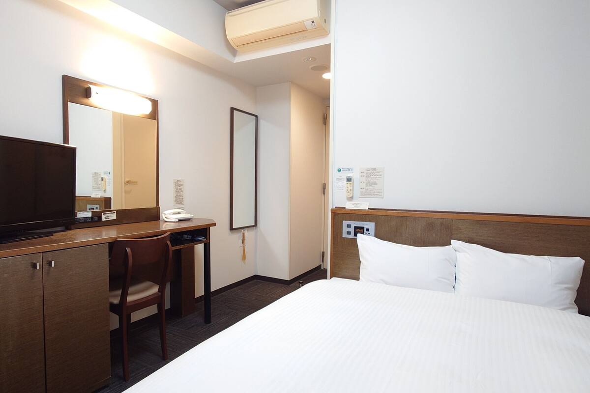 A hotel room with a bed, a desk, a chair, a television, a mirror, and a wall-mounted phone.