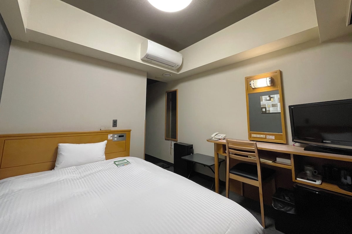 A hotel room with a bed, a desk, a chair, a television, a mirror, and an air conditioning unit.
