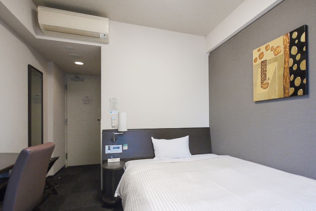 A hotel room with a bed, a desk, a chair, a bedside table, a wall-mounted air conditioner, and a painting.