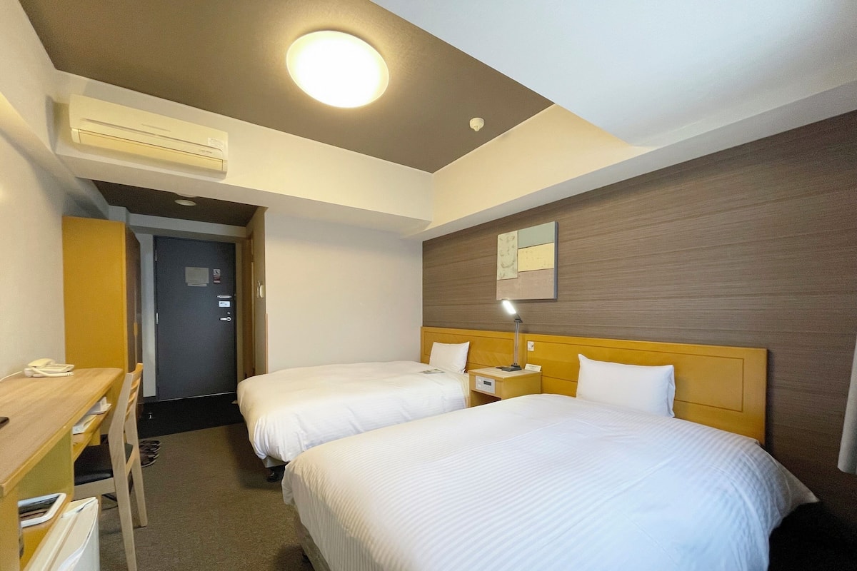 A hotel room with two beds, a desk, a chair, an air conditioning unit, and a bedside table with a lamp.