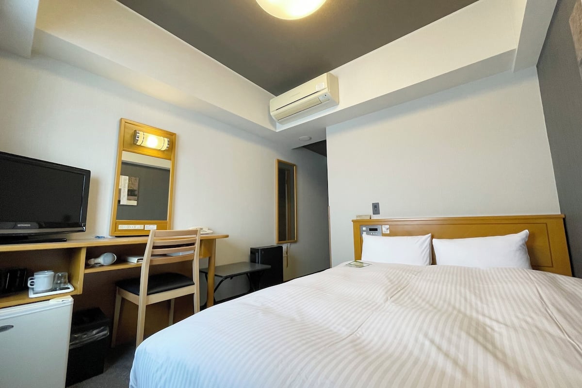 A hotel room with a bed, a desk with a chair, a television, and an air conditioning unit.