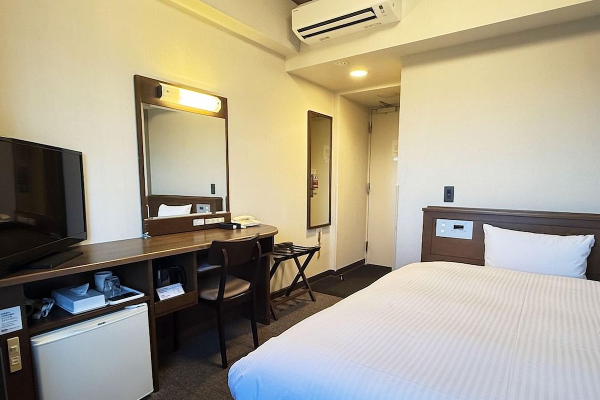 A hotel room with a bed, a desk with a chair, a television, a mini-fridge, a mirror, and an air conditioning unit.