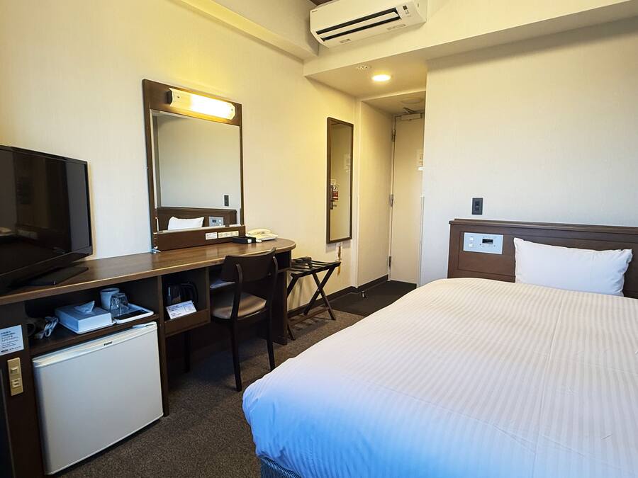 A hotel room with a bed, a desk with a chair, a television, a mirror, and a mini-fridge.