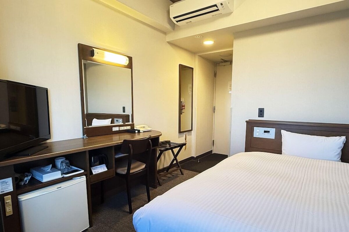 A hotel room with a bed, a desk with a chair, a television, a mini-fridge, a mirror, and an air conditioning unit.