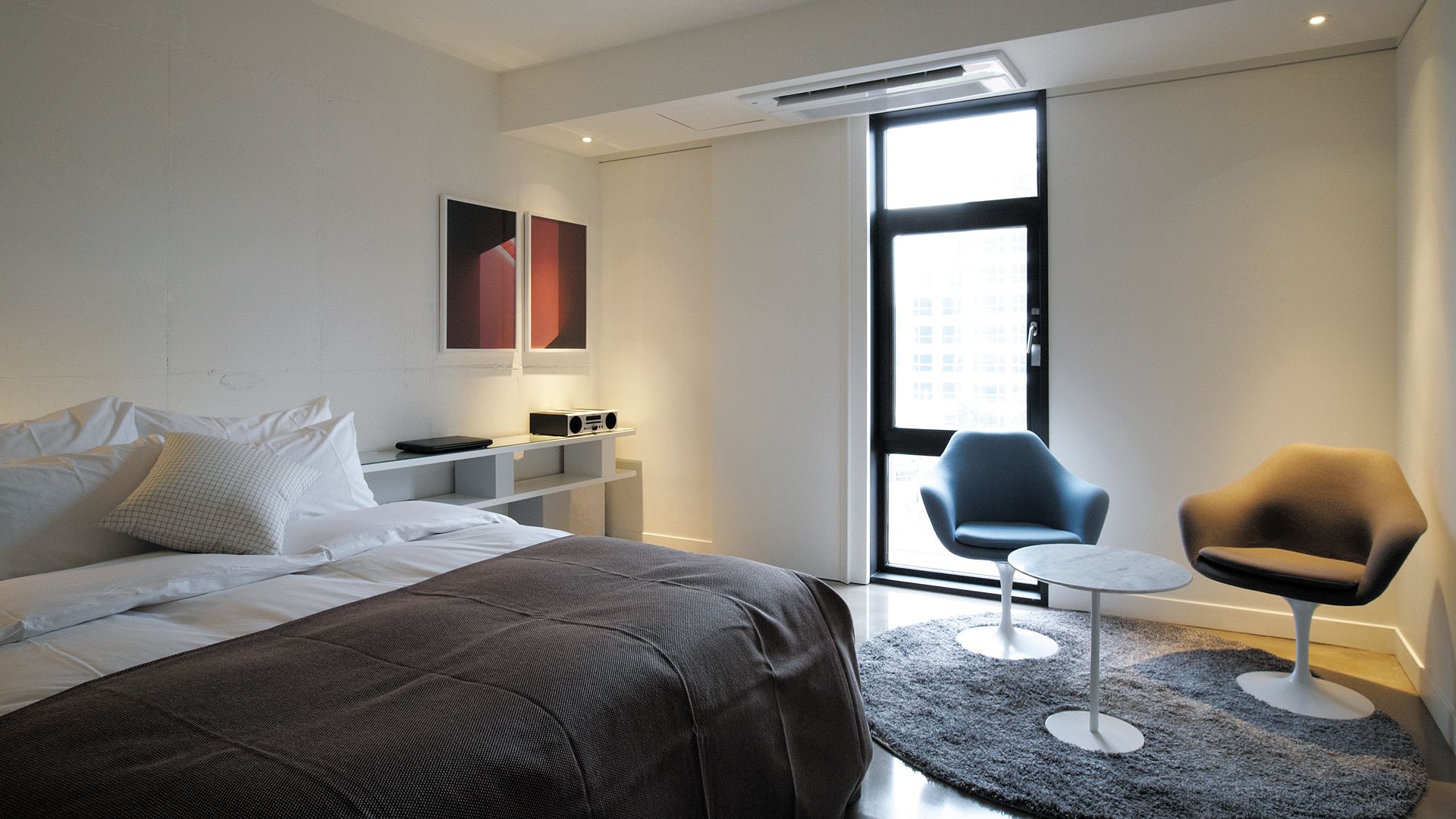 A modern hotel room with a large bed, a small desk, a chair, and a view through a large window.