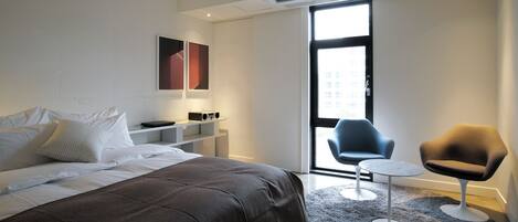 A modern hotel room with a large bed, a small desk, a chair, and a view through a large window.