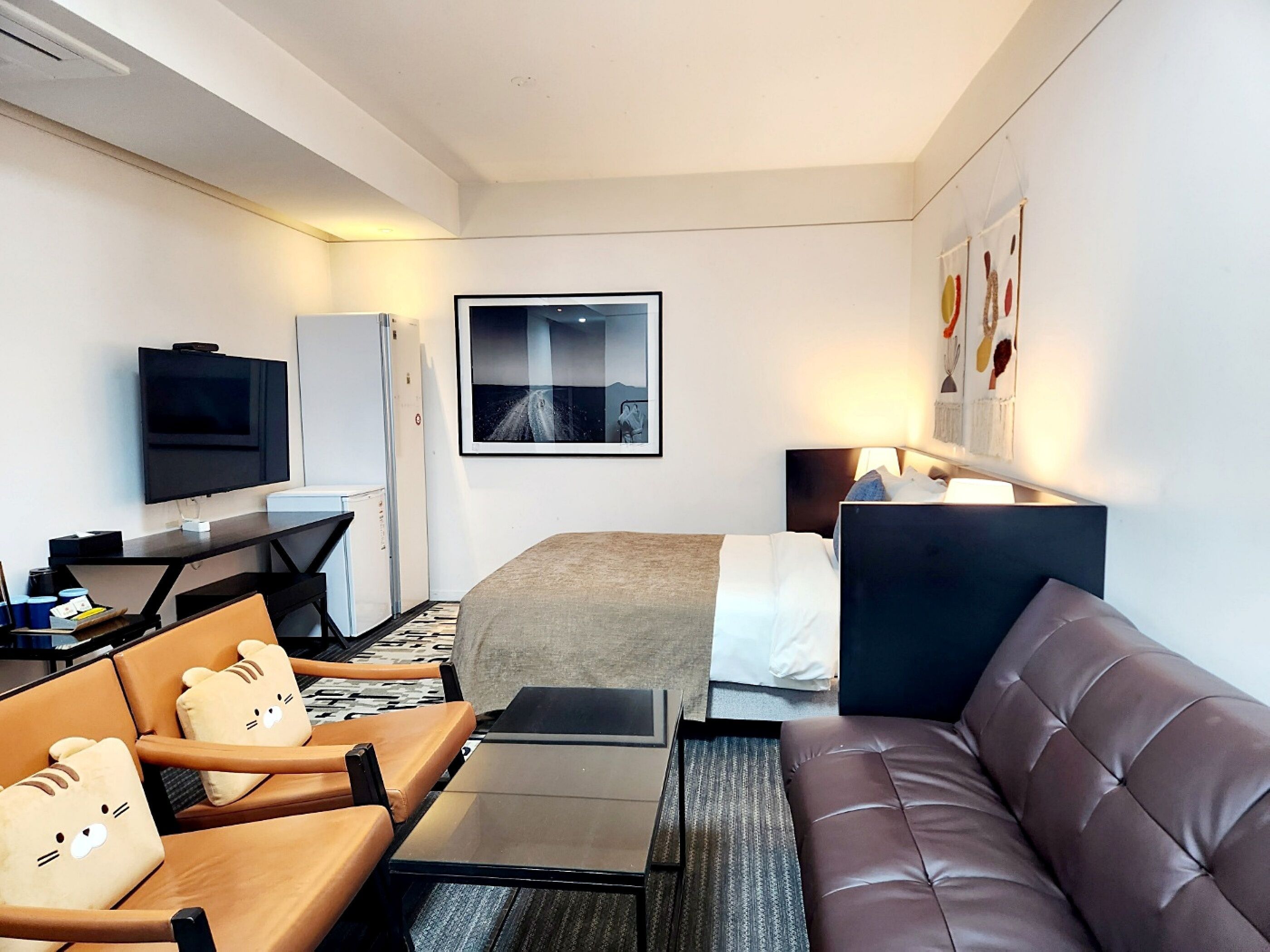 A modern hotel room with a bed, a sofa, a TV, a desk, and a chair with bear-themed cushions.