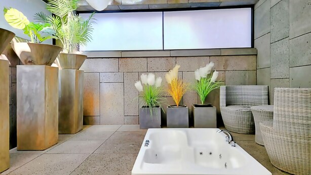 Un espacio exterior moderno con un jacuzzi grande de color blanco, plantas en macetas y mobiliario de mimbre.