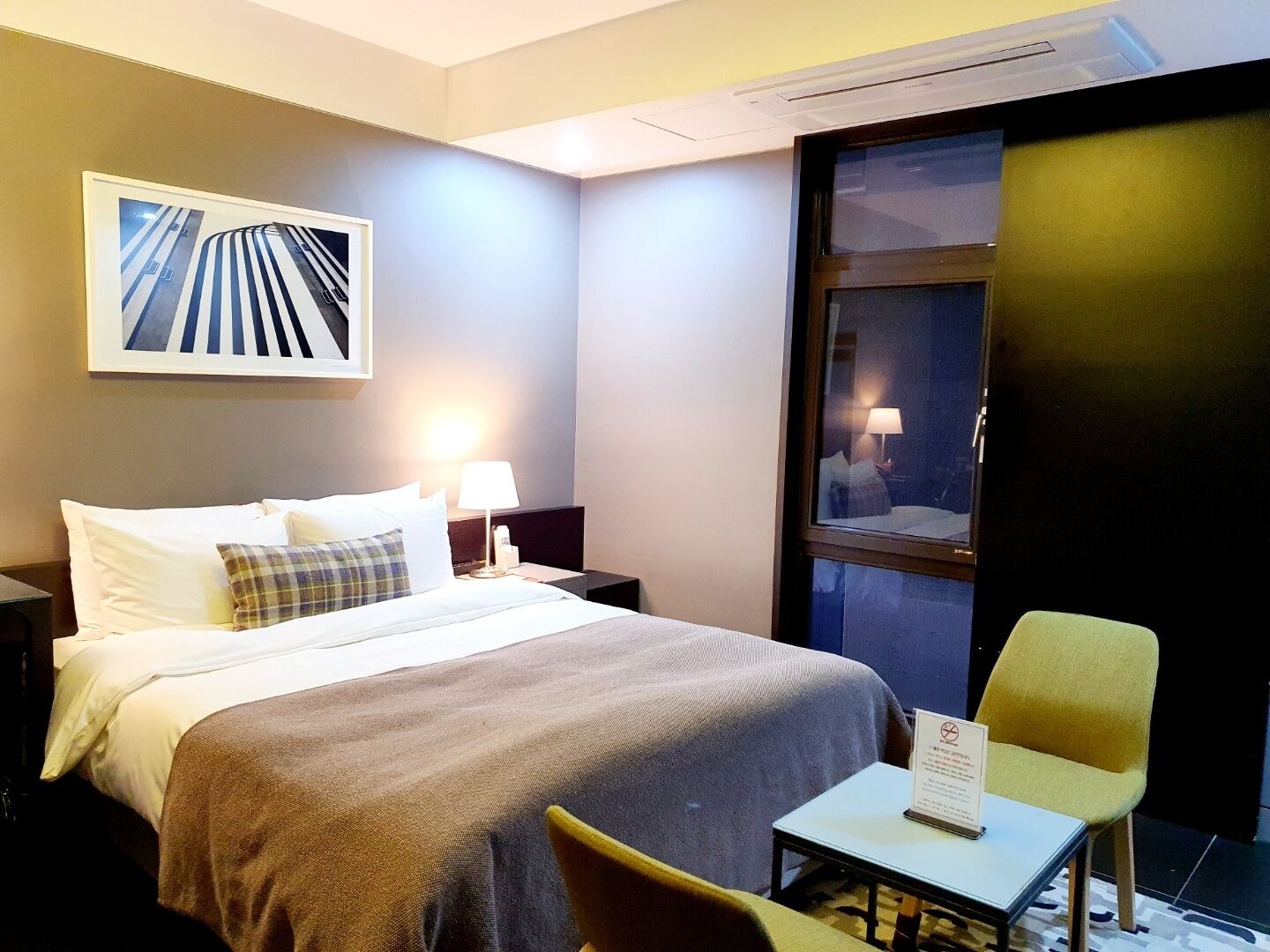 Una habitación de hotel con cama, mesita de noche, silla, pequeña mesa y ventana.