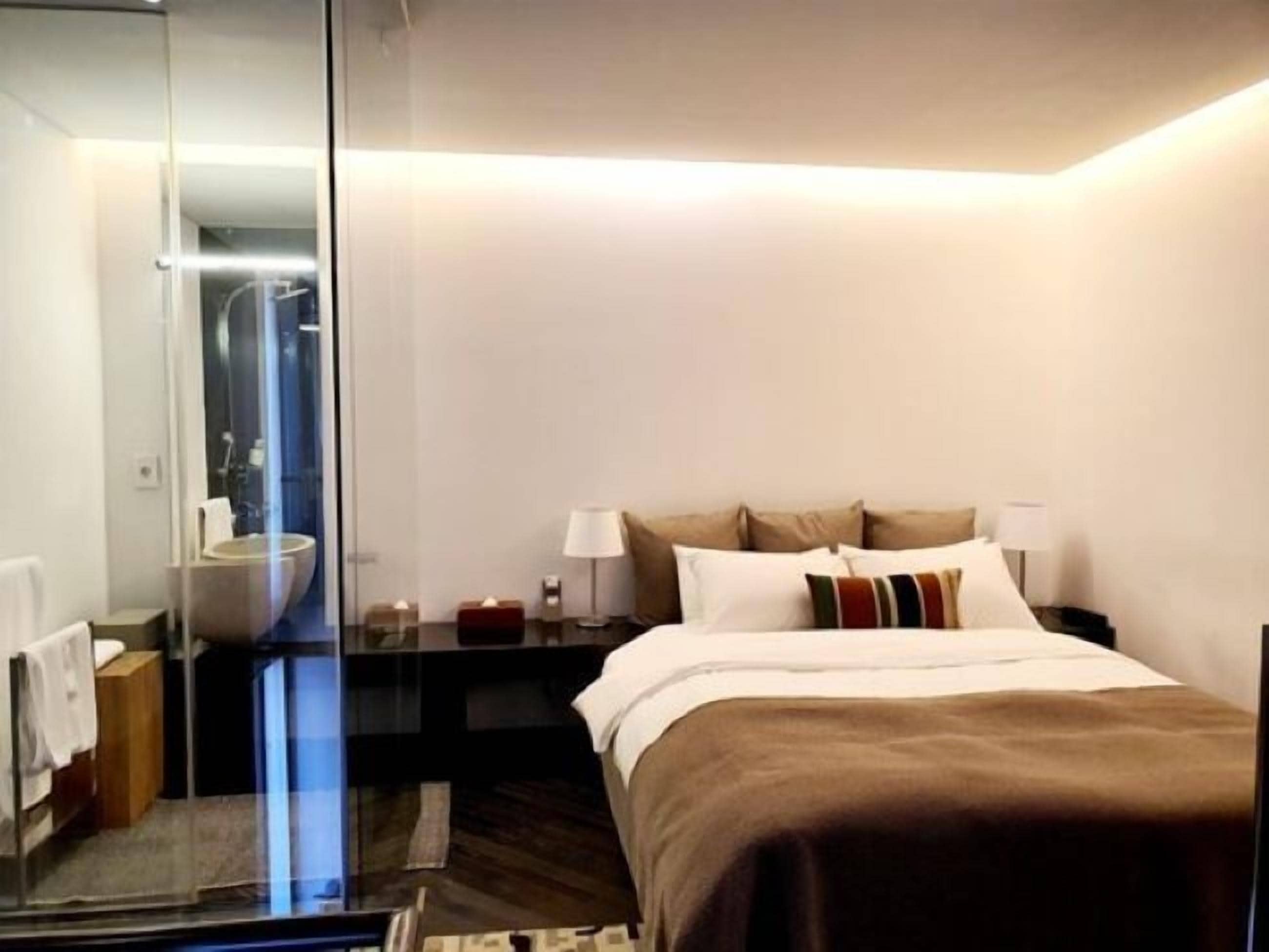 Habitación de hotel moderna con una cama grande, mesitas de noche, un espejo y un separador de vidrio que divide el baño.
