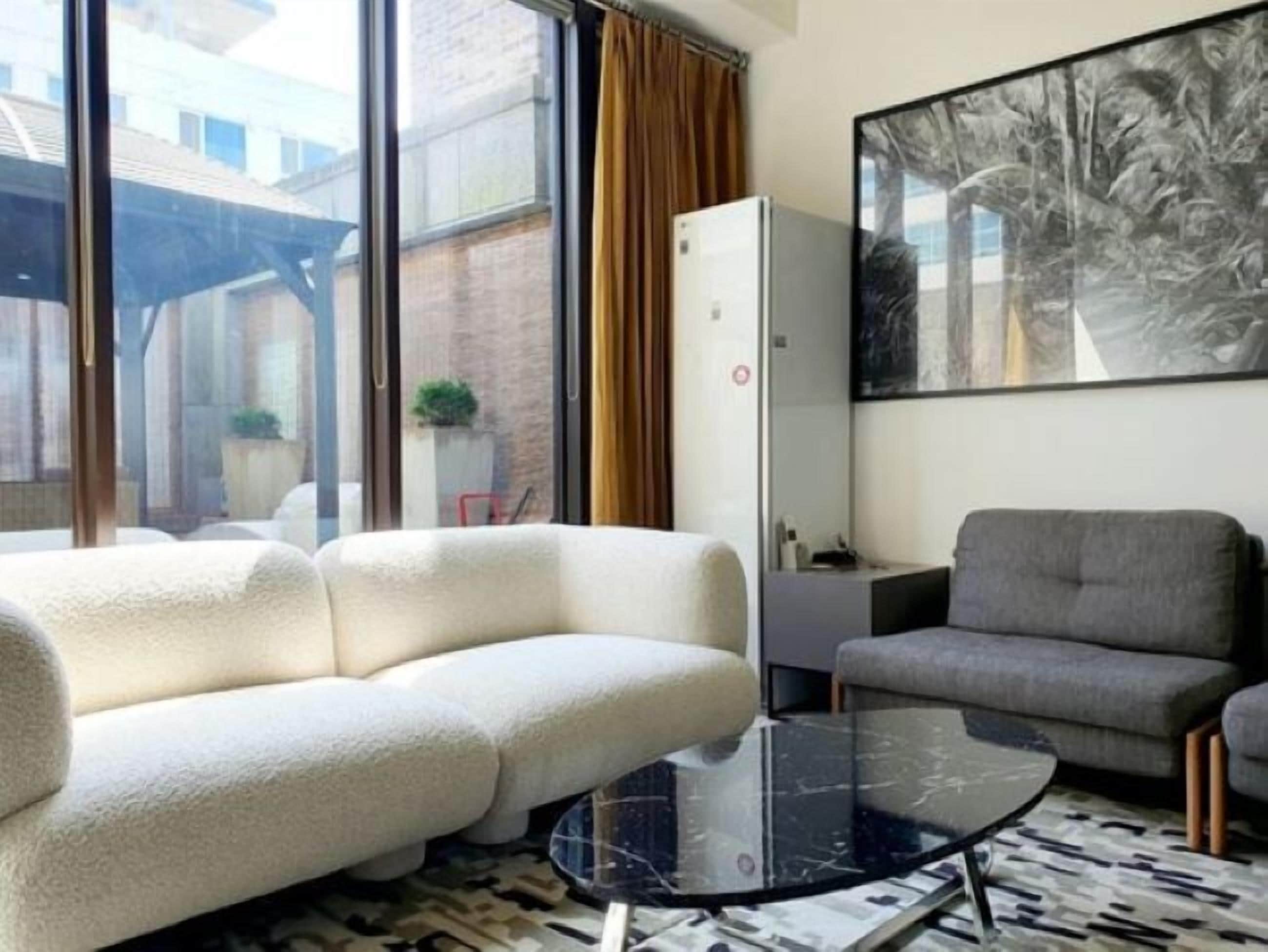 Un salon moderne avec un canapé blanc, un fauteuil gris et une table basse en verre. Une grande fenêtre est agrémentée de rideaux, un tableau orne le mur et une petite plante est posée sur une table.
