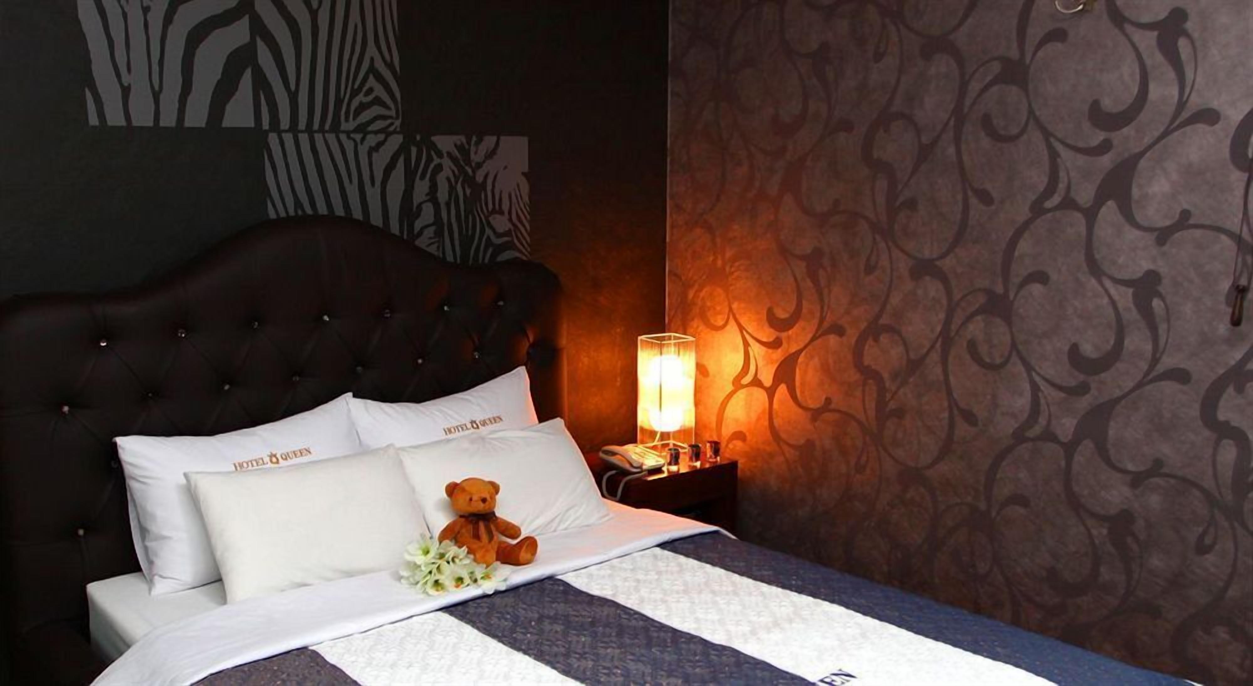 A hotel room with a tufted headboard, a bed with white bedding, a teddy bear, and a nightstand with a lamp.